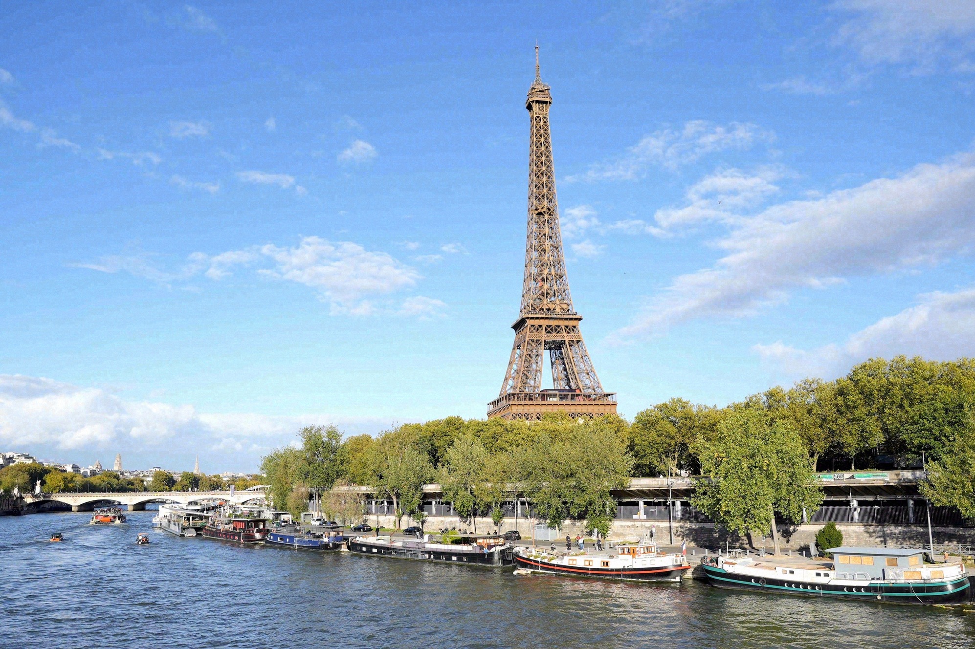 Eiffel-torni kesällä Seineltä kuvattuna, vedessä risteilyveneitä ja keskietäisyydellä silta, taivaalla sinistä ja kesäpilviä.