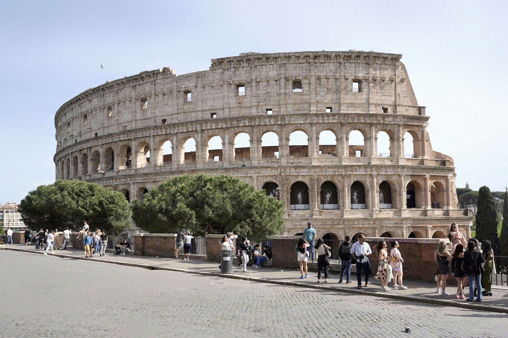 Colosseum kuvattuna kivetyltä Piazza del Colosseolta, ympärillä matkailijoita ja reunoilla pinjoja.